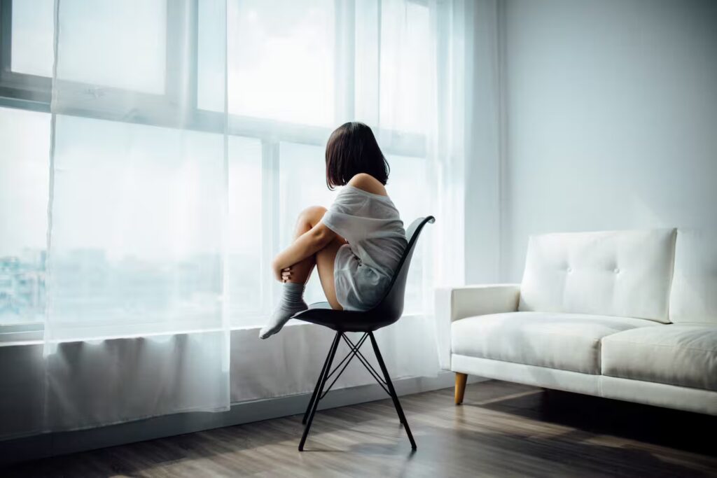 Woman sitting on a chair looking outside, struggling to balance coping mechanisms and actual growth.
